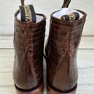 May include: A pair of brown leather boots with a crocodile skin texture. The boots have a pull tab with the word "CARRAJE" in gold lettering. The soles are a lighter brown color.