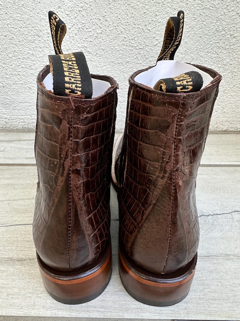 May include: A pair of brown leather boots with a crocodile skin texture. The boots have a pull tab with the word "CARRAJE" in gold lettering. The soles are a lighter brown color.