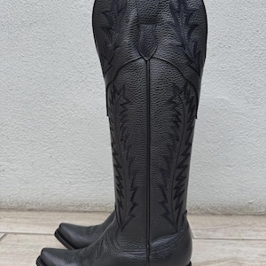 Pode incluir: Um par de botas de cowboy pretas com costuras decorativas. As botas t&ecirc;m bico fino e apar&ecirc;ncia de couro texturizado. A haste alta apresenta desenhos bordados intrincados. As botas est&atilde;o em uma superf&iacute;cie de madeira.