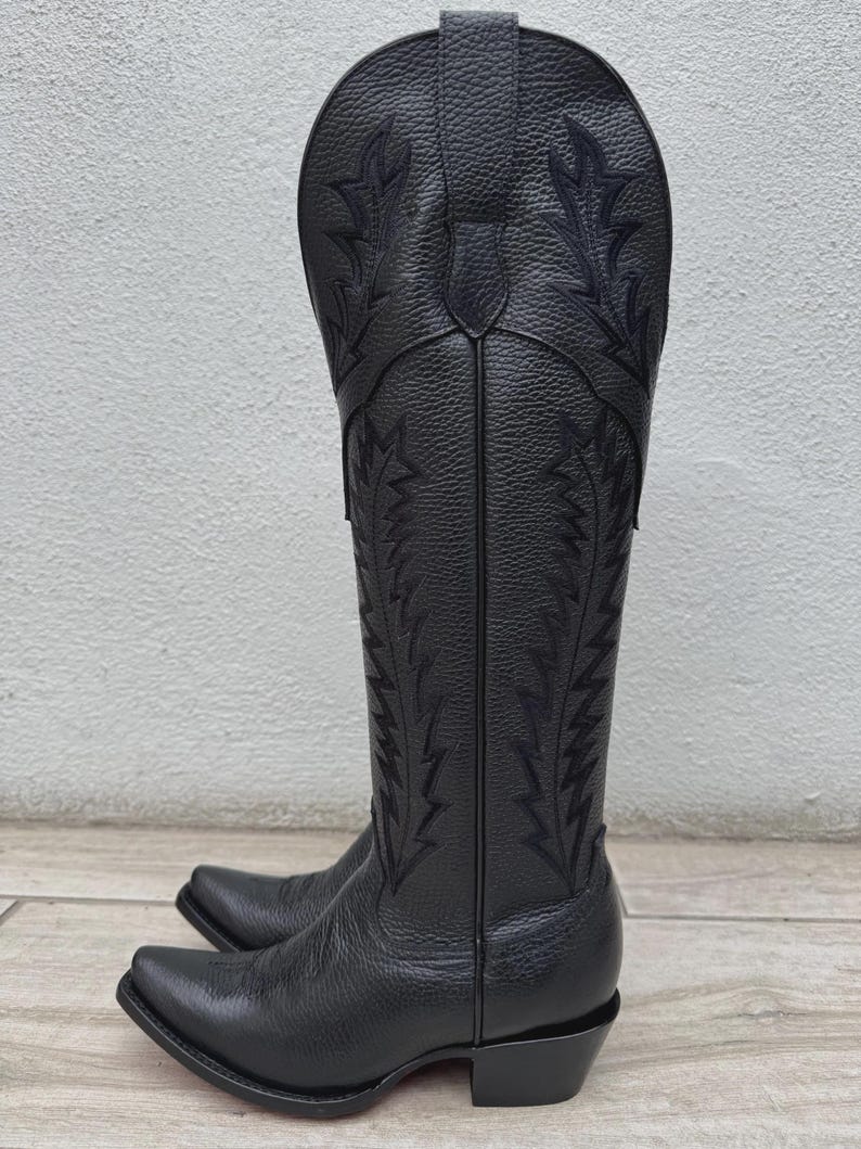 Pode incluir: Um par de botas de cowboy pretas com costuras decorativas. As botas t&ecirc;m bico fino e apar&ecirc;ncia de couro texturizado. A haste alta apresenta desenhos bordados intrincados. As botas est&atilde;o em uma superf&iacute;cie de madeira.
