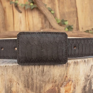 May include: A black belt with a textured, rectangular buckle and matching strap. The belt is made of a dark material with a subtle pattern, and is displayed on a wooden surface.