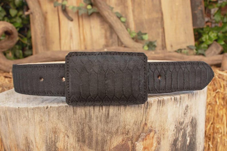 May include: A black belt with a textured, rectangular buckle and matching strap. The belt is made of a dark material with a subtle pattern, and is displayed on a wooden surface.