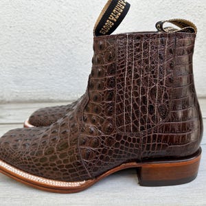 May include: Brown leather cowboy boots with a square toe and a textured, crocodile-like pattern. The boots have a pull tab at the top and a stacked heel. The sole is trimmed with a light-colored stitching.