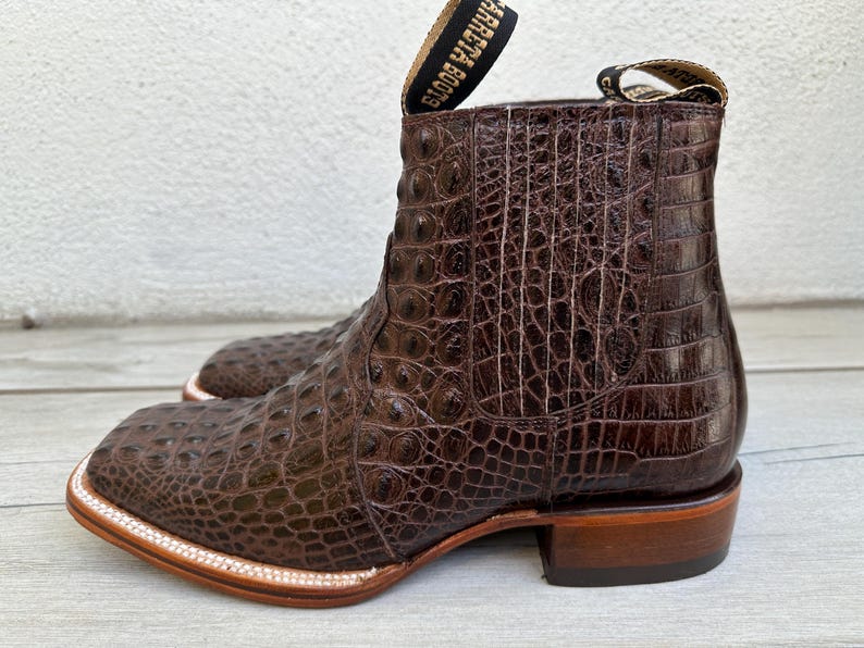 May include: Brown leather cowboy boots with a square toe and a textured, crocodile-like pattern. The boots have a pull tab at the top and a stacked heel. The sole is trimmed with a light-colored stitching.