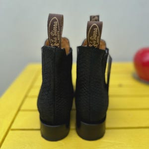 May include: Black ankle boots with a textured design. Each boot features a pull tab with the "La Sierra Boots" logo. The boots are displayed on a yellow surface.