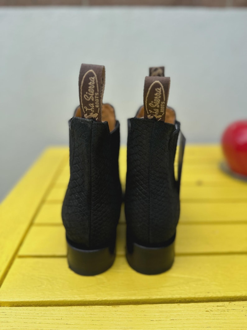 May include: Black ankle boots with a textured design. Each boot features a pull tab with the "La Sierra Boots" logo. The boots are displayed on a yellow surface.