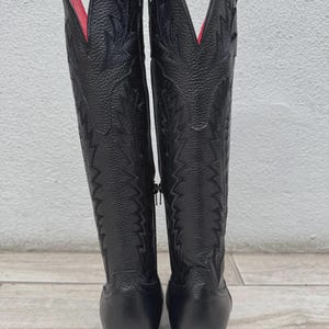 Pode incluir: Um par de botas de cowboy de couro preto com costuras decorativas e forro interior vermelho. As botas t&ecirc;m bico fino e salto empilhado. Um z&iacute;per percorre a lateral de cada bota.