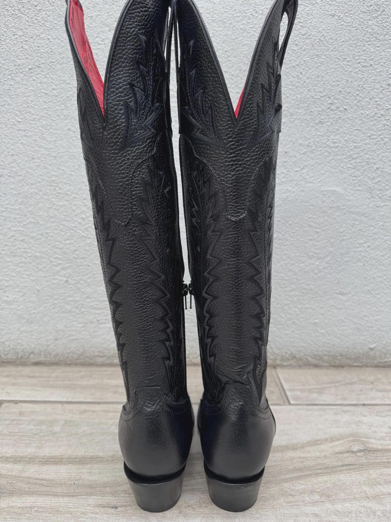 Pode incluir: Um par de botas de cowboy de couro preto com costuras decorativas e forro interior vermelho. As botas t&ecirc;m bico fino e salto empilhado. Um z&iacute;per percorre a lateral de cada bota.