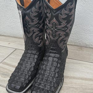 Puede incluir: Un par de botas de vaquero negras con punta cuadrada y un patrón texturizado similar al de un cocodrilo. Las botas presentan costuras decorativas rojas y blancas en la caña superior y un borde cosido blanco alrededor de la suela.