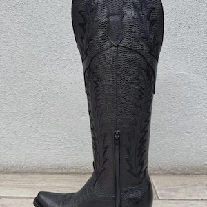 Pode incluir: Bota de cowboy em couro preto com costuras decorativas e z&iacute;per lateral. A bota tem bico fino e salto empilhado. A bota tem aproximadamente 35.5 cm de altura.
