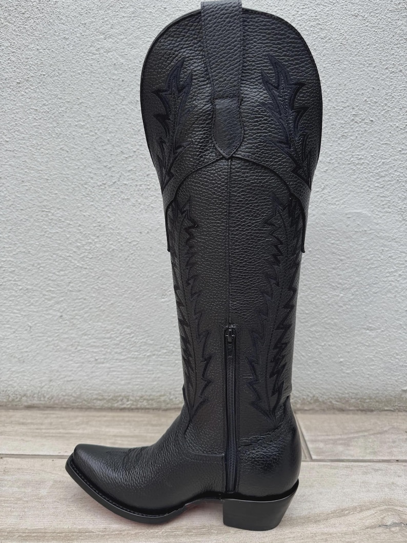 Pode incluir: Bota de cowboy em couro preto com costuras decorativas e z&iacute;per lateral. A bota tem bico fino e salto empilhado. A bota tem aproximadamente 35.5 cm de altura.
