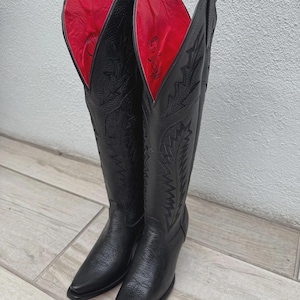 Pode incluir: Um par de botas de cowboy de couro preto com costuras intrincadas e bico pontudo. O forro interior &eacute; vermelho vibrante. As botas s&atilde;o altas, atingindo a metade da panturrilha, e t&ecirc;m um pequeno salto.