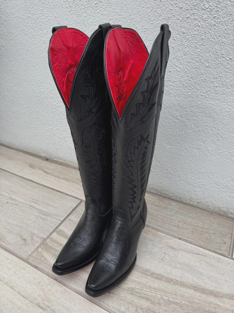 Pode incluir: Um par de botas de cowboy de couro preto com costuras intrincadas e bico pontudo. O forro interior &eacute; vermelho vibrante. As botas s&atilde;o altas, atingindo a metade da panturrilha, e t&ecirc;m um pequeno salto.