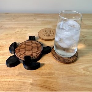 May include: Wooden turtle-shaped coaster set with a glass of water. The set includes a turtle-shaped coaster, a round coaster with the text "ATOM ADAM", and a coaster with a honeycomb pattern. The turtle coaster is black with a brown, honeycomb-patterned shell.