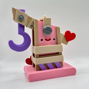 May include: A whimsical, 3D-printed sculpture featuring a pink rectangular base, a pink and tan body, and a purple hook. The sculpture has a smiling face and red heart accents. The design incorporates geometric shapes and a playful aesthetic.