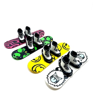 May include: Four miniature snowboards with attached boots. Designs include a pink and black swirl, green alien heads on black, yellow smiley faces, and a white board with a monster face.