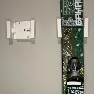 May include: A pair of white wall-mounted ski holders next to a green and white patterned ski. The ski has a black binding and the word "SHARP" in white. The ski has a bandana-style pattern.