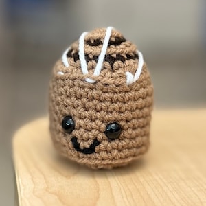 Peut inclure: Une petite figurine amigurumi crochetée en fil marron clair. Elle présente une partie supérieure marron foncé avec des détails en fil blanc, deux yeux de sécurité en plastique noir et un petit sourire brodé en noir sur la face avant.