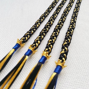Puede incluir: Cuatro llaveros trenzados negros y dorados con borlas azules y dijes de corona dorada.