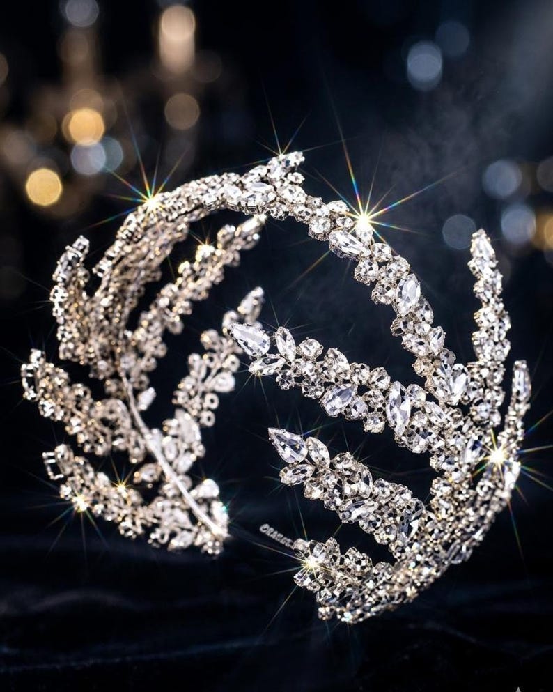 K&ouml;nnte beinhalten: Eine funkelnde silberne Tiara mit einem Blatt- und Blumenmuster. Die Tiara ist mit zahlreichen klaren Kristallen verziert, die das Licht reflektieren. Das Design weist mehrere B&auml;nder und eine geschwungene Form auf, ideal f&uuml;r besondere Anl&auml;sse.