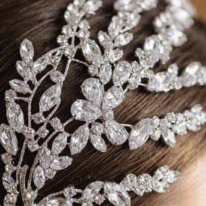 K&ouml;nnte beinhalten: Ein silberfarbenes Braut-Haarst&uuml;ck mit klaren, funkelnden Kristallen. Das Haarst&uuml;ck hat ein Blumen- und Bl&auml;tterdesign mit gro&szlig;en, tropfenf&ouml;rmigen Kristallen und kleineren, runden Steinen. Das filigrane Design ist vor einem dunklen Hintergrund angeordnet.