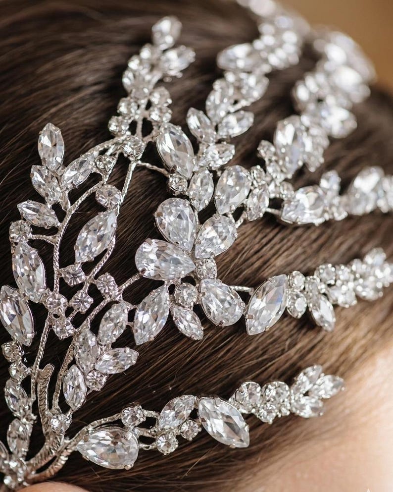 K&ouml;nnte beinhalten: Ein silberfarbenes Braut-Haarst&uuml;ck mit klaren, funkelnden Kristallen. Das Haarst&uuml;ck hat ein Blumen- und Bl&auml;tterdesign mit gro&szlig;en, tropfenf&ouml;rmigen Kristallen und kleineren, runden Steinen. Das filigrane Design ist vor einem dunklen Hintergrund angeordnet.