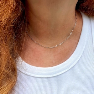 May include: A delicate silver chain necklace with a paperclip link design. The necklace is worn on a person wearing a white ribbed tank top. The person has long, wavy auburn hair.