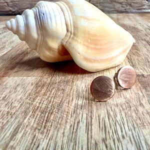 May include: A pair of copper-colored, round stud earrings with a brushed metal finish. The earrings are displayed on a wooden surface next to a large, cream-colored seashell. The earrings are a simple, minimalist design.