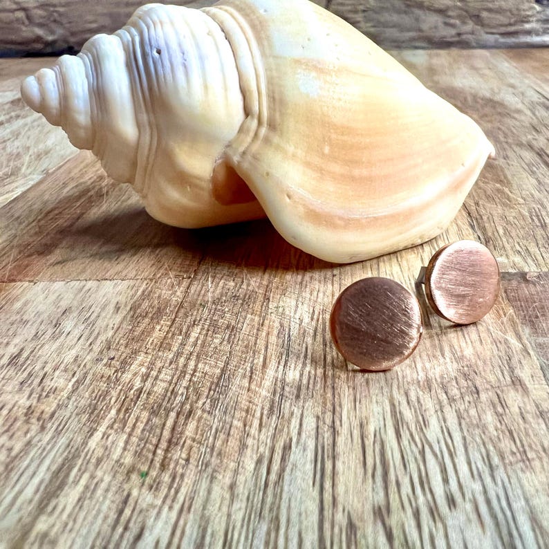 May include: A pair of copper-colored, round stud earrings with a brushed metal finish. The earrings are displayed on a wooden surface next to a large, cream-colored seashell. The earrings are a simple, minimalist design.