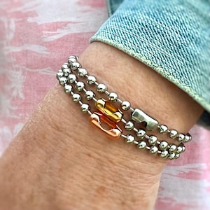 May include: A silver chain bracelet with three rows of beads and two gold and rose gold accent pieces.