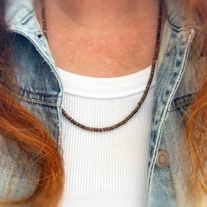 May include: A brown beaded necklace worn over a white t-shirt and a denim jacket.