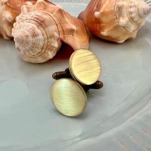 May include: A pair of gold-colored cufflinks with an oval design. The cufflinks have a brushed metal finish and a dark brown base. The background includes seashells on a light blue surface.