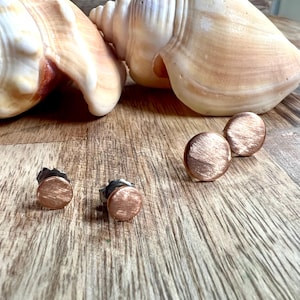 May include: A pair of copper-colored stud earrings with a brushed metal finish. The earrings are round and sit on silver posts. Two seashells are in the background on a wooden surface.