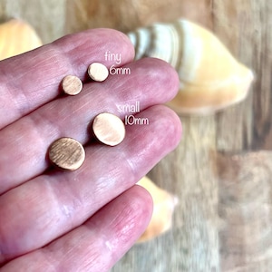 May include: A hand displays four small, round, copper-colored stud earrings. The text "tiny 6mm" and "small 10mm" are next to the earrings. The earrings are on a wooden surface with a seashell in the background.