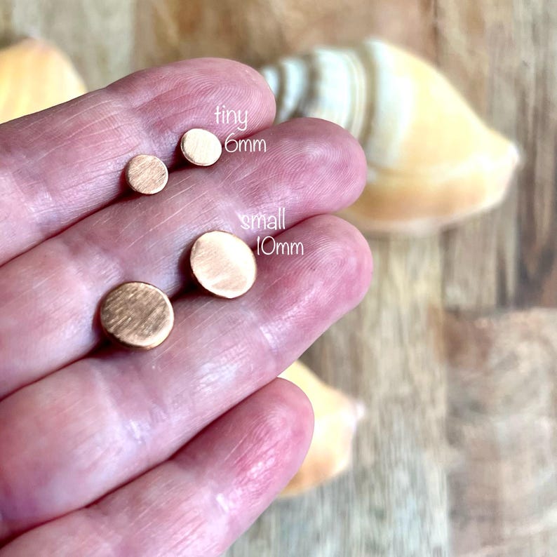 May include: A hand displays four small, round, copper-colored stud earrings. The text "tiny 6mm" and "small 10mm" are next to the earrings. The earrings are on a wooden surface with a seashell in the background.