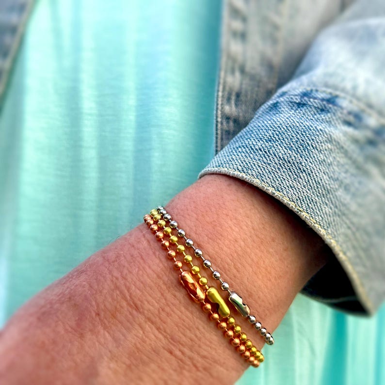 May include: Three bracelets stacked on a person's wrist. The top bracelet is silver, the middle bracelet is gold, and the bottom bracelet is copper.