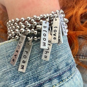 May include: A silver chain bracelet with multiple charms. The charms are rectangular and have words engraved on them, including "love", "bloom", "happy", "Aloha", "bloom", and "brave".