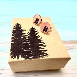 May include: A pair of copper cufflinks with a tree design, presented in a brown box with a tree silhouette printed on the lid.