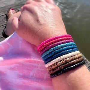 May include: A stack of colorful beaded bracelets in shades of pink, blue, brown, and black. The bracelets are made of round wooden beads and are stacked on a person's wrist.