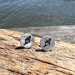 May include: Two silver-colored stud earrings with a black seashell design. The earrings are round and have a brushed metal finish. The background shows a wooden surface and the ocean.