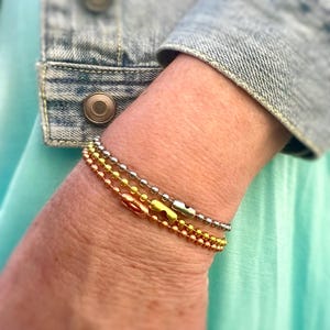 May include: Three layered bracelets with silver, gold, and rose gold chain links. The bracelets are stacked on a person's wrist.