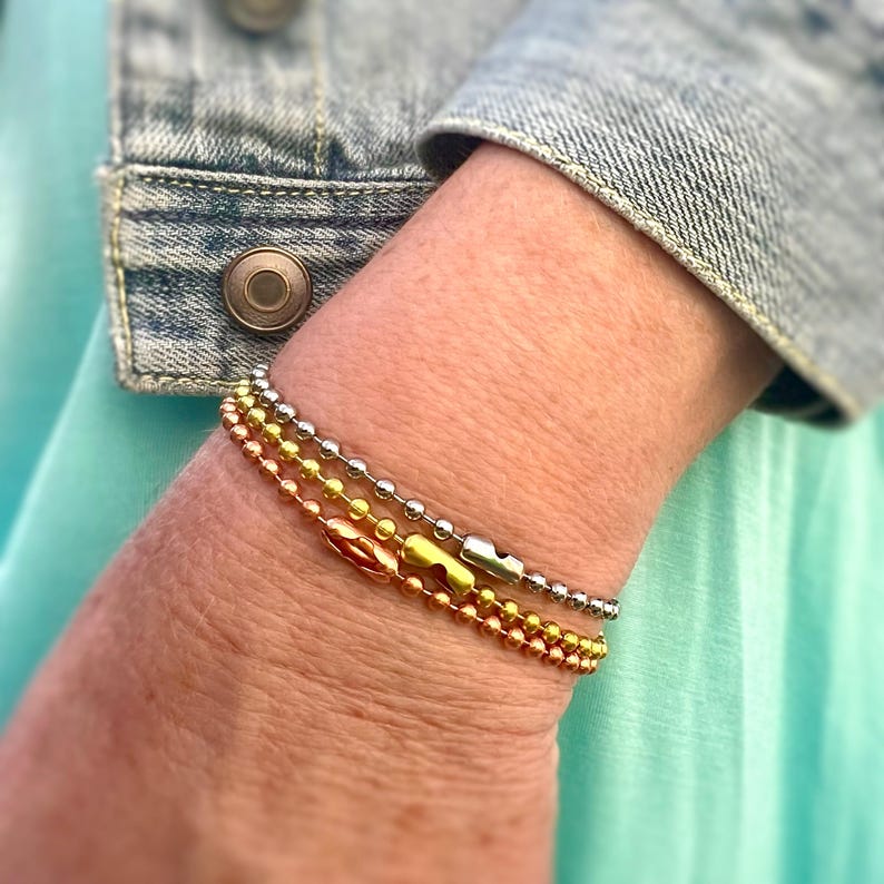 May include: Three layered bracelets with silver, gold, and rose gold chain links. The bracelets are stacked on a person's wrist.