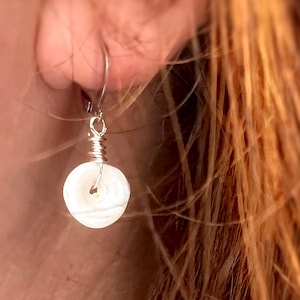 May include: A single drop earring featuring a small, circular, white shell bead suspended from a silver-toned metal wire hook. The bead is attached to the hook with a coiled silver-toned wire wrap that secures the shell through its center hole.