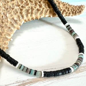 May include: Black beaded necklace with alternating sections of black, white, and pastel-colored beads. The necklace is displayed on a white wooden surface with a starfish in the background. The necklace is a casual accessory.
