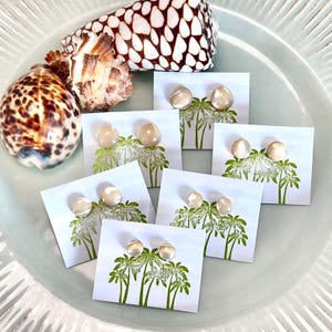 May include: A collection of pearl stud earrings displayed on cards with a palm tree design. Two seashells are placed near the earrings on a light blue plate. The earrings have a light, pearlescent color.