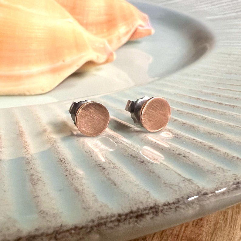 May include: A pair of small, round stud earrings with a brushed copper finish. The earrings are set against a light blue, ridged plate, with a seashell in the background. The earrings have a simple, minimalist design.