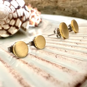 May include: Gold-colored, round stud earrings with a brushed finish. The earrings are displayed on a white and brown textured surface, with a seashell in the background. The earrings have a simple, minimalist design.