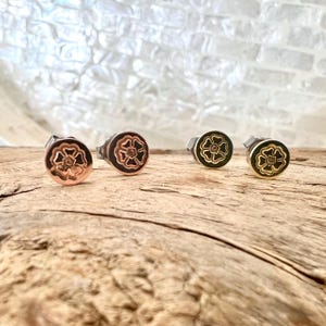 May include: Four pairs of stud earrings with a floral design. The earrings are in various colors, including copper, black, and gold. The earrings are displayed on a wooden surface.