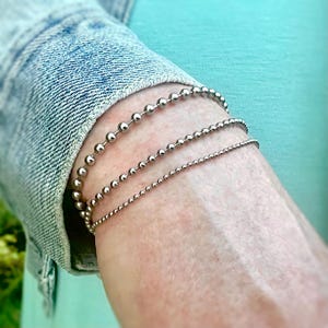 May include: Three silver beaded bracelets are worn on a wrist. The bracelets vary in bead size, with the top bracelet having the largest beads. The wrist is against a light teal background and a denim jacket.