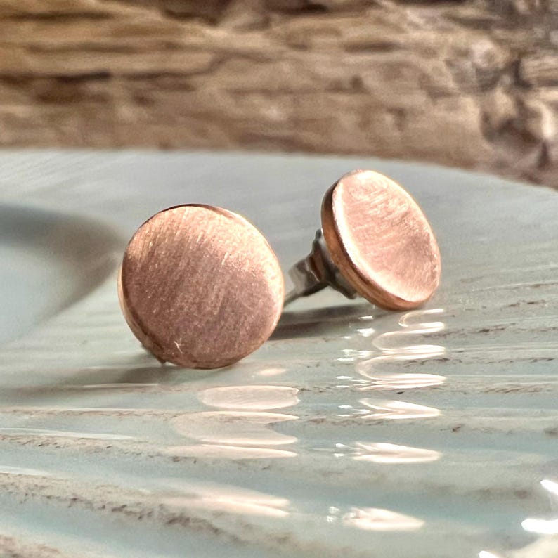 May include: A pair of copper-colored stud earrings. Each earring features a circular, flat design with a brushed metal texture. The earrings are set on silver-colored posts, showcasing a minimalist aesthetic.